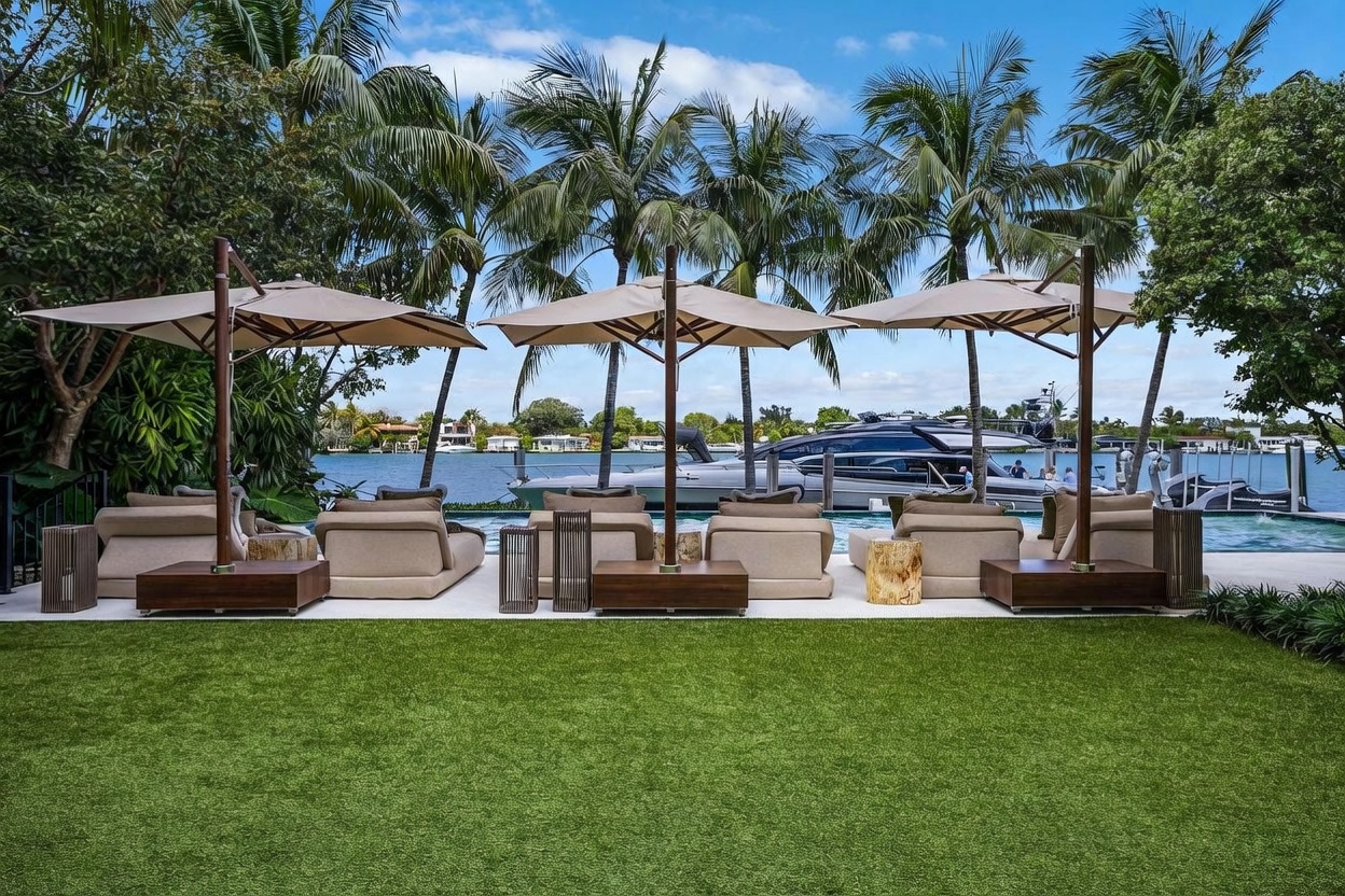 This luxurious outdoor space features a pristine infinity-edge pool overlooking a waterfront canal with a docked yacht. The patio area is furnished with plush lounge chairs shaded by large, modern umbrellas, set against a backdrop of lush palm trees and manicured green lawn. The scene captures a serene, high-end resort-style atmosphere perfect for relaxation and entertaining.