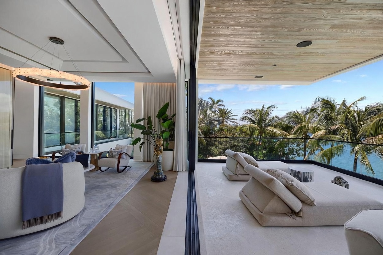 This sophisticated living area seamlessly transitions to an expansive outdoor balcony through large, retractable glass walls. The interior features light wood flooring, a modern circular chandelier, and plush seating, while the balcony offers comfortable lounge chairs overlooking a lush, tropical waterfront view. The space emphasizes an open-concept design that blurs the line between indoor luxury and the natural beauty of the exterior landscape.