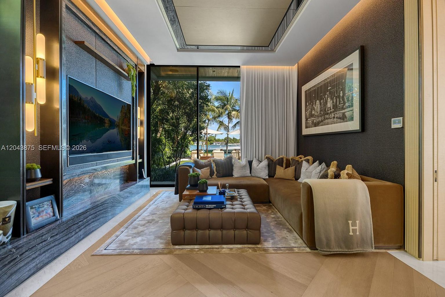 This sophisticated living room features a plush, L-shaped brown sofa centered around a large, tufted leather ottoman on a textured area rug. A wall-mounted television is integrated into a dark, textured feature wall with built-in shelving and vertical lighting accents, while floor-to-ceiling glass doors provide a seamless view of the lush outdoor greenery and waterfront. The space is finished with light wood flooring and a framed art piece, creating a warm, luxurious atmosphere.