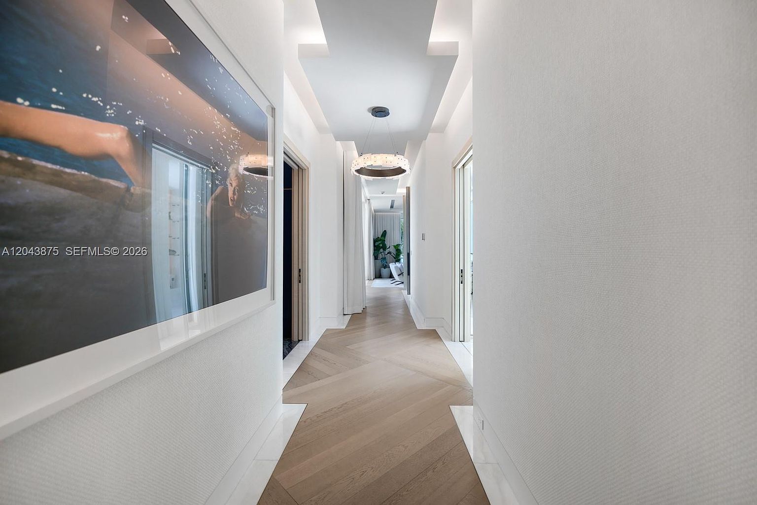 This elegant hallway features light-toned wood flooring arranged in a chevron pattern, bordered by crisp white marble trim. A large, framed photograph hangs on the left wall, while a modern, circular chandelier with a textured finish provides a focal point from the recessed ceiling. The perspective looks down the bright, minimalist corridor toward a sunlit room at the far end, creating a sense of depth and sophisticated flow.