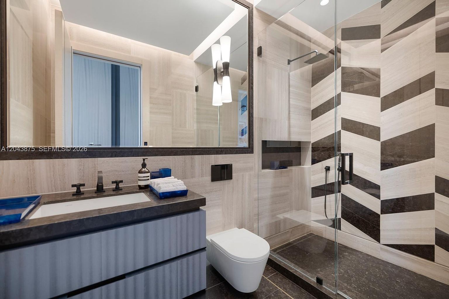 This modern bathroom features a sleek vanity with a stone countertop, a wall-mounted toilet, and a walk-in shower enclosed by glass panels. The shower area is a focal point, showcasing a striking chevron-patterned tile wall in contrasting dark and light tones. The space is illuminated by a contemporary light fixture mounted on a large mirror, creating a sophisticated and high-end aesthetic.