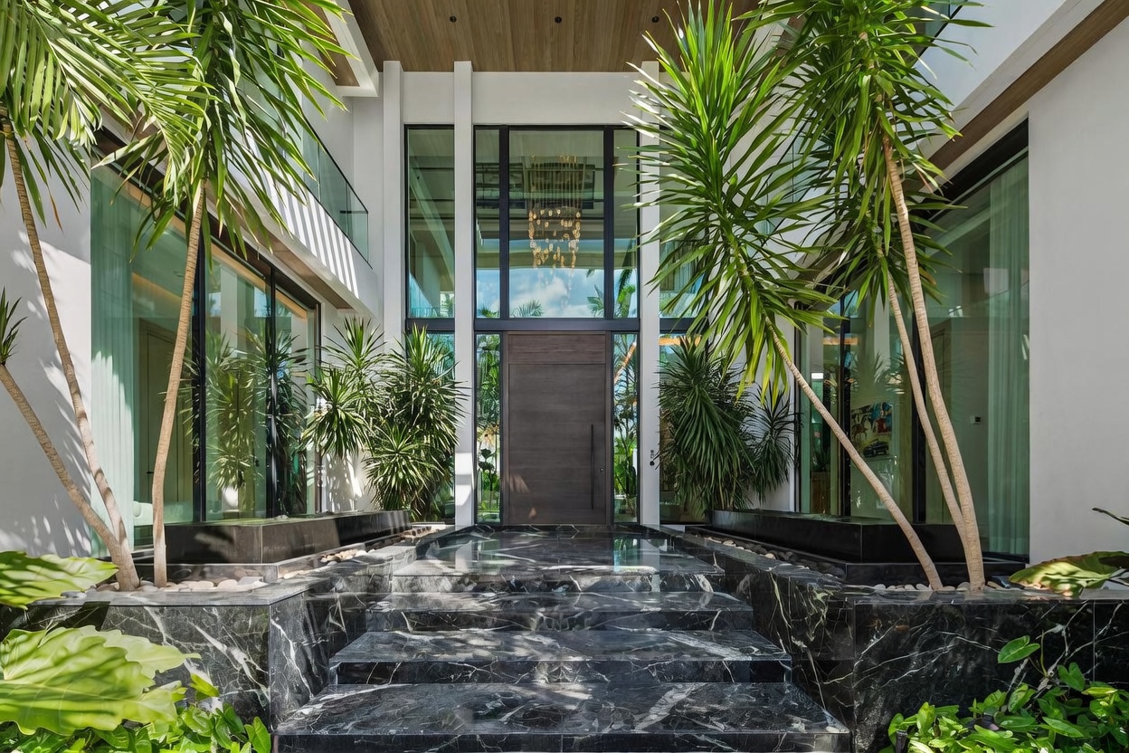 This grand entryway features a dramatic set of dark stone steps leading up to a massive, modern wooden front door. The entrance is framed by tall, floor-to-ceiling glass panels and lush tropical palm trees, creating a sophisticated and welcoming atmosphere. The symmetrical design and reflective stone surfaces contribute to an impression of high-end luxury and architectural elegance.