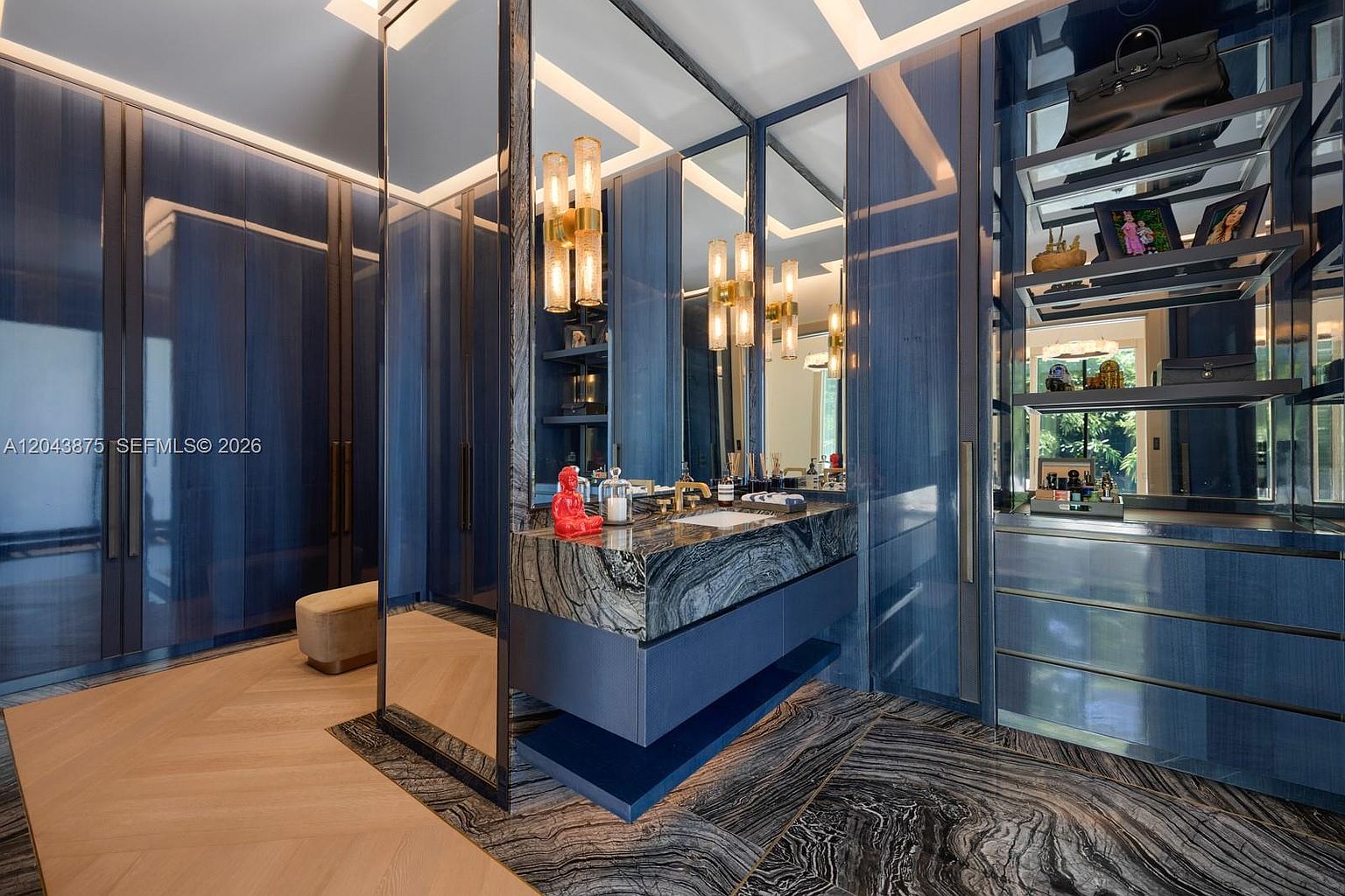 This luxurious walk-in closet features high-gloss blue cabinetry and a central vanity area with a striking stone countertop and integrated sink. The space is accented by warm, vertical light fixtures and a large mirror, while the flooring transitions between light wood planks and a dramatic, dark patterned stone. The overall aesthetic is sophisticated and modern, offering a high-end dressing experience.