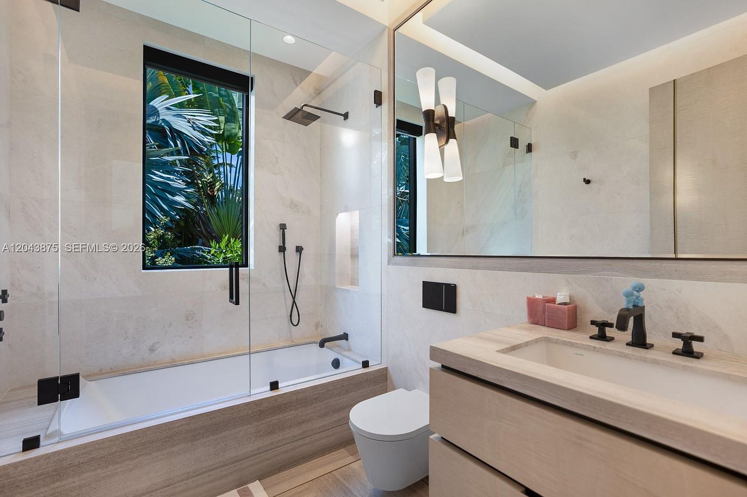 This modern bathroom features a sleek vanity with a light wood finish and a dark faucet, paired with a wall-mounted toilet. A glass-enclosed shower and tub combination is positioned against a window that looks out onto lush greenery, creating a bright and serene atmosphere. The space is finished with neutral stone tiles and a large, minimalist mirror, offering a clean and contemporary aesthetic.