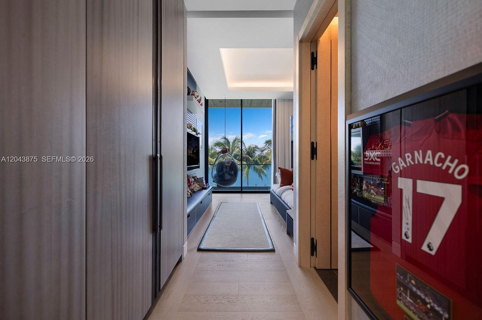 This modern bedroom features a sleek, narrow layout with light wood flooring and a built-in storage unit along the left wall. A hanging chair is positioned near the large glass doors that provide a clear view of palm trees and the water beyond. To the right, a framed sports jersey is displayed on the wall, adding a personalized touch to the contemporary space.