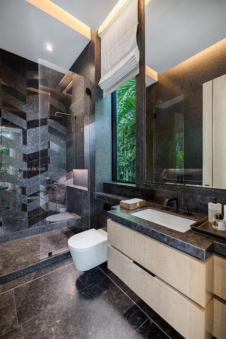 This modern bathroom features a striking shower enclosure with a chevron-patterned dark stone wall and a glass partition. A floating light-wood vanity with a dark stone countertop sits beneath a large mirror, while a wall-mounted toilet is positioned next to a tall window overlooking lush greenery. The space is defined by its sophisticated, moody aesthetic and high-end finishes.