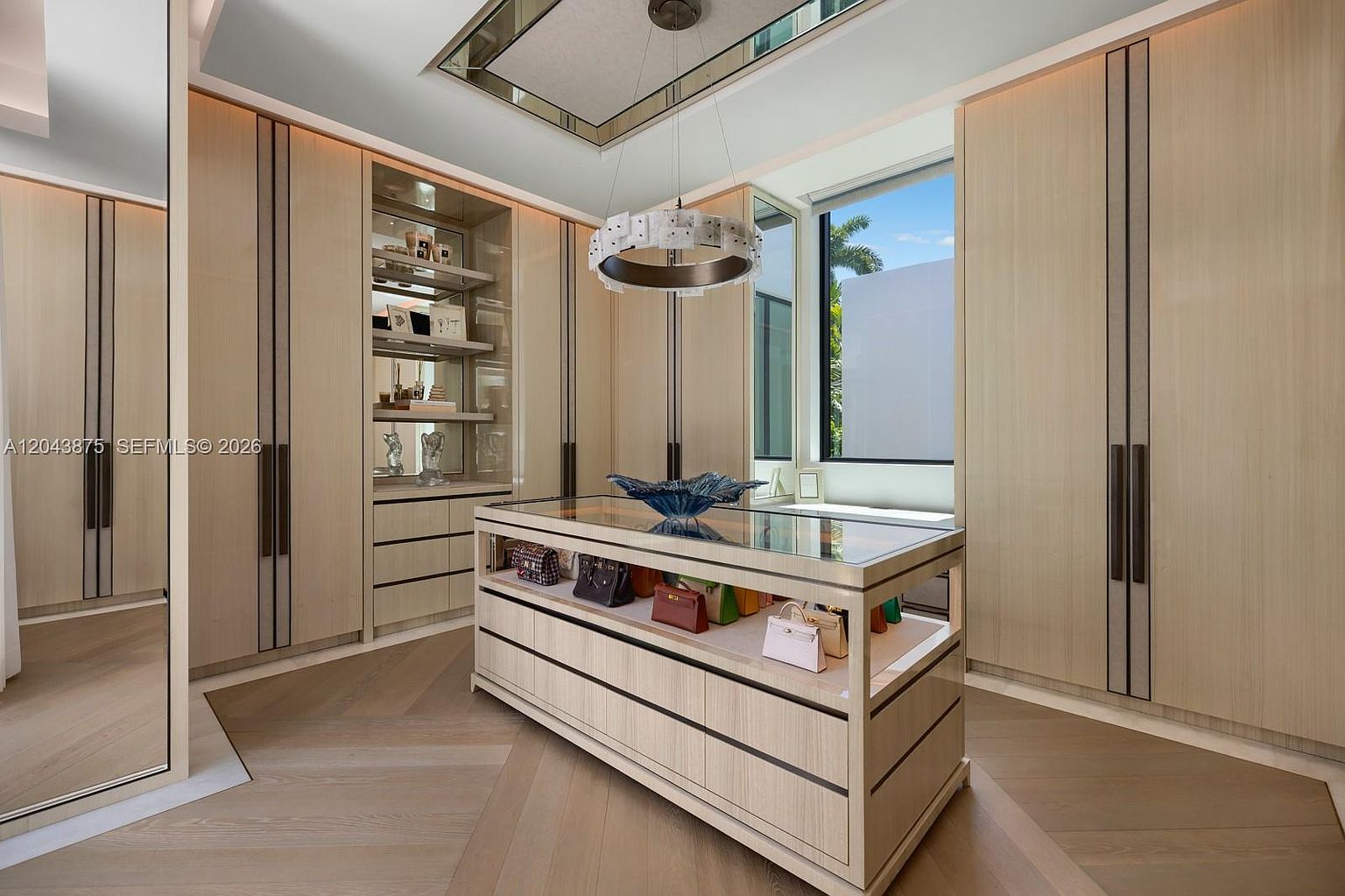 This luxurious walk-in closet features custom light wood cabinetry with integrated shelving and a central island topped with a glass display surface. A unique circular chandelier hangs from the ceiling, while a large window provides natural light and a view of the outdoors. The space is organized with high-end finishes, including a mirrored wall and light-toned wood flooring, creating an elegant and sophisticated atmosphere.