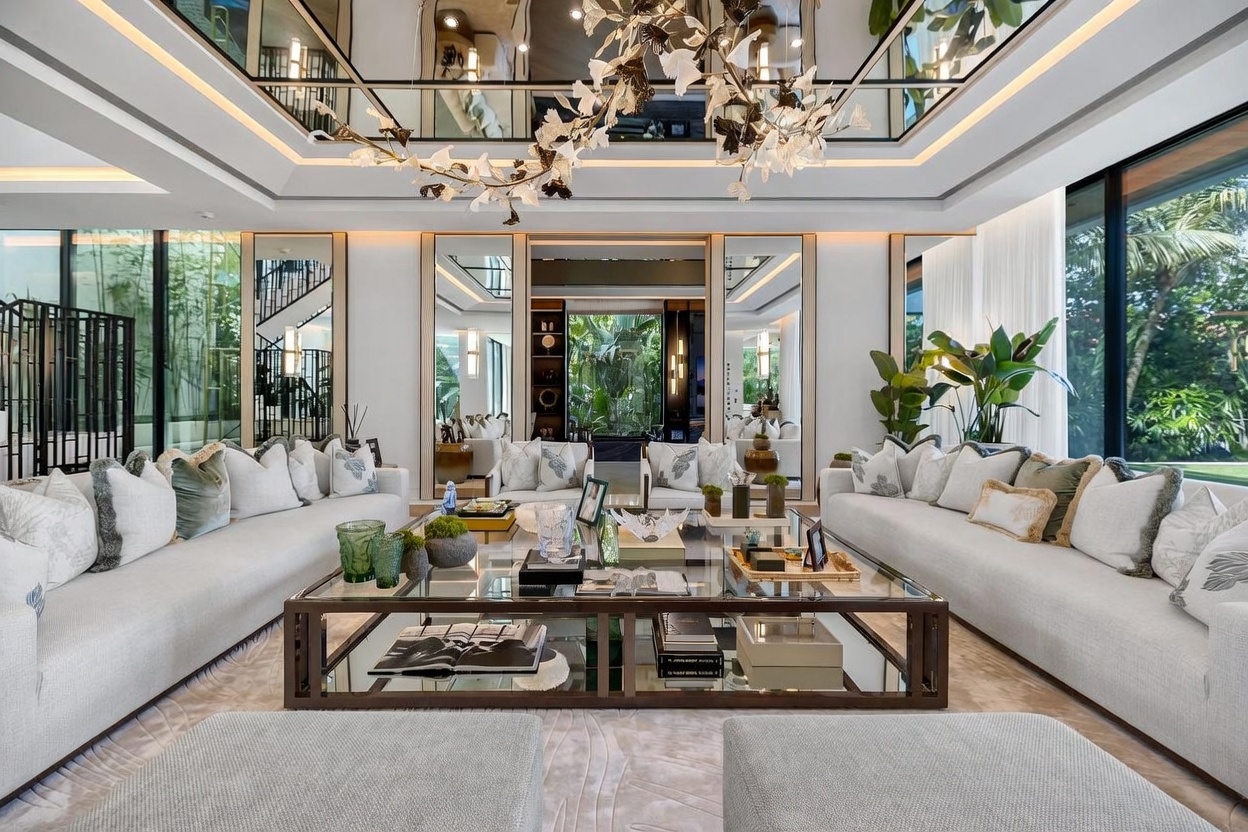 This luxurious living room features two large, light-colored sofas facing a central glass-topped coffee table, creating a symmetrical and sophisticated seating arrangement. The space is defined by a mirrored ceiling with integrated lighting, tall wall mirrors that reflect the lush greenery outside, and a striking, sculptural chandelier. Large windows provide an expansive view of the outdoor landscape, enhancing the bright and airy atmosphere of this high-end interior.