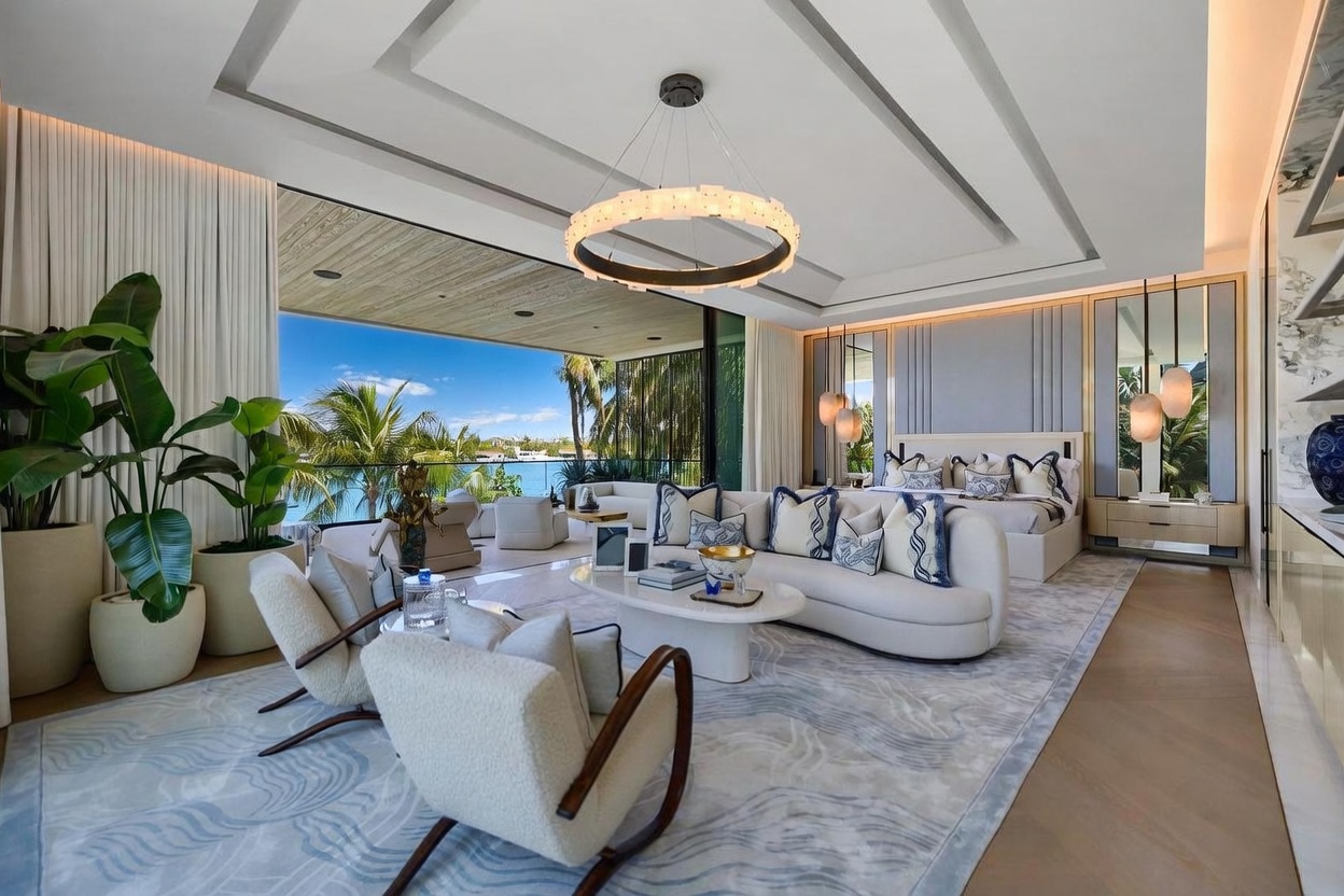 This expansive primary bedroom suite features a seamless transition to a covered outdoor terrace through large glass sliding doors, offering panoramic water views. The room is elegantly furnished with a curved sofa, plush armchairs, and a king-sized bed, all anchored by a large, patterned area rug. A sophisticated circular chandelier serves as a focal point, while the neutral color palette and clean lines create a serene, high-end atmosphere.