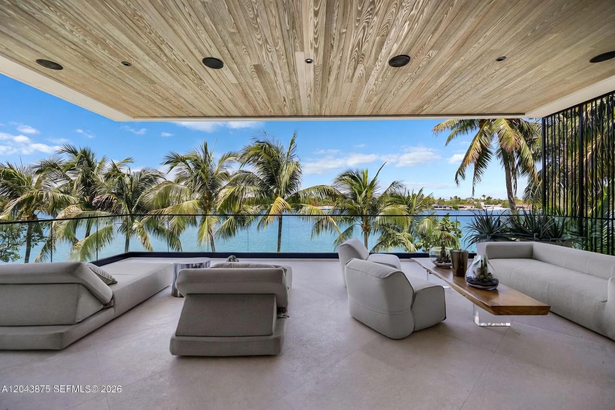 This expansive, covered balcony offers a luxurious outdoor living space with a panoramic view of the water and palm trees. The area is furnished with contemporary, low-profile lounge seating and a sleek wooden coffee table, all set against a backdrop of floor-to-ceiling glass railings. The ceiling features warm-toned wood paneling with recessed lighting, creating a sophisticated and serene atmosphere perfect for relaxation.