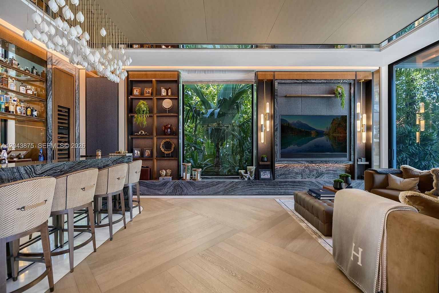 This sophisticated living space features a bar area with a stone-topped counter and high-backed stools, complemented by a large television mounted on a dark, textured wall. A central window provides a lush, green view of the outdoors, while light wood flooring in a herringbone pattern adds warmth to the room. The space is elegantly furnished with a plush brown sofa and a low-profile ottoman, creating a luxurious and inviting atmosphere.