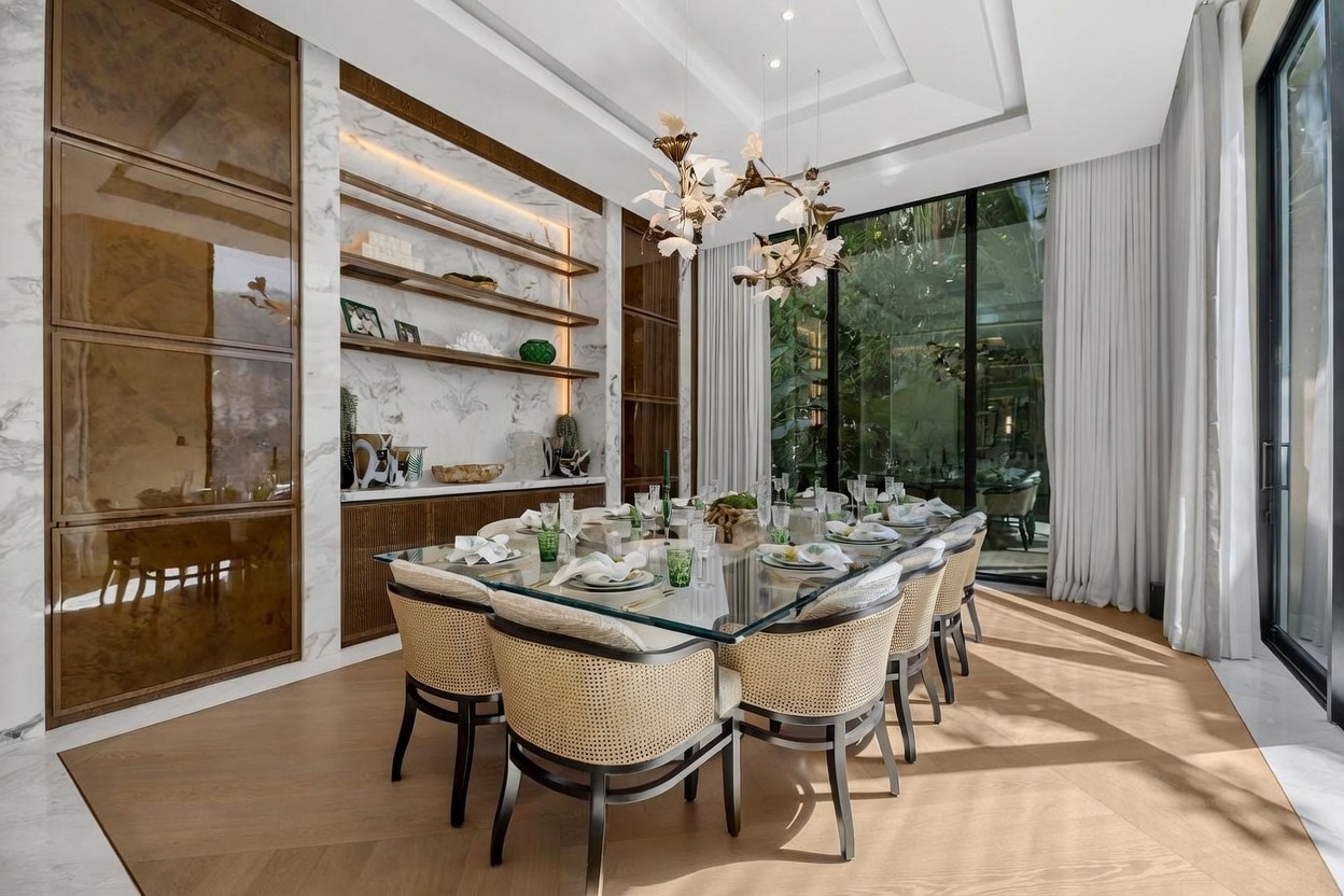 This elegant dining room features a large glass-topped table surrounded by chairs with woven cane backs and light-toned upholstery. The space is anchored by a wall with built-in shelving and cabinetry set against a stone-patterned backdrop, while floor-to-ceiling windows provide an expansive view of the lush greenery outside. A sculptural, nature-inspired chandelier hangs above the table, casting a sophisticated glow over the light wood flooring.
