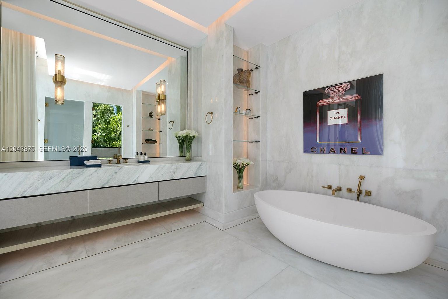 This luxurious primary bathroom features a sleek, freestanding white soaking tub positioned next to a large wall-mounted art piece. A long vanity with a stone countertop and integrated sink sits beneath a massive mirror, complemented by modern gold-toned fixtures and warm accent lighting. The space is finished with light-toned stone surfaces and includes built-in glass shelving, creating a bright and sophisticated atmosphere.