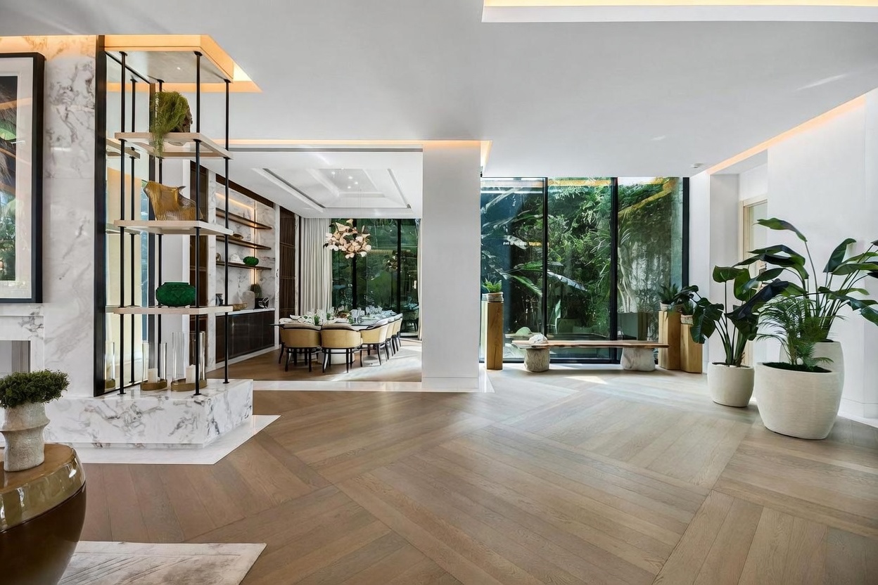 This expansive, open-concept living area features light wood flooring laid in a chevron pattern and a striking, multi-tiered display shelf with a stone base. The space flows seamlessly into a formal dining room and is illuminated by natural light from large windows that look out onto a lush, green garden. The interior design is sophisticated and modern, characterized by clean lines, neutral tones, and a sense of airy, architectural elegance.