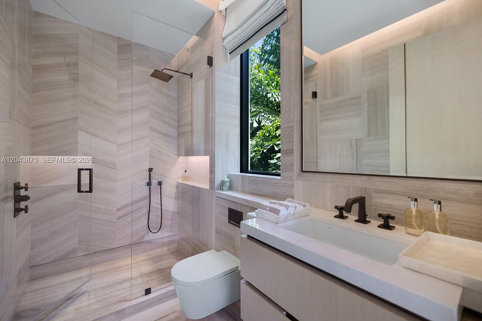 This modern bathroom features a seamless glass-enclosed walk-in shower with chevron-patterned stone wall tiles and a rainfall showerhead. A floating vanity with a white countertop and dark hardware sits beneath a large mirror, while a wall-mounted toilet is positioned next to a tall window overlooking lush greenery. The space is characterized by its neutral color palette, clean lines, and sophisticated, spa-like atmosphere.