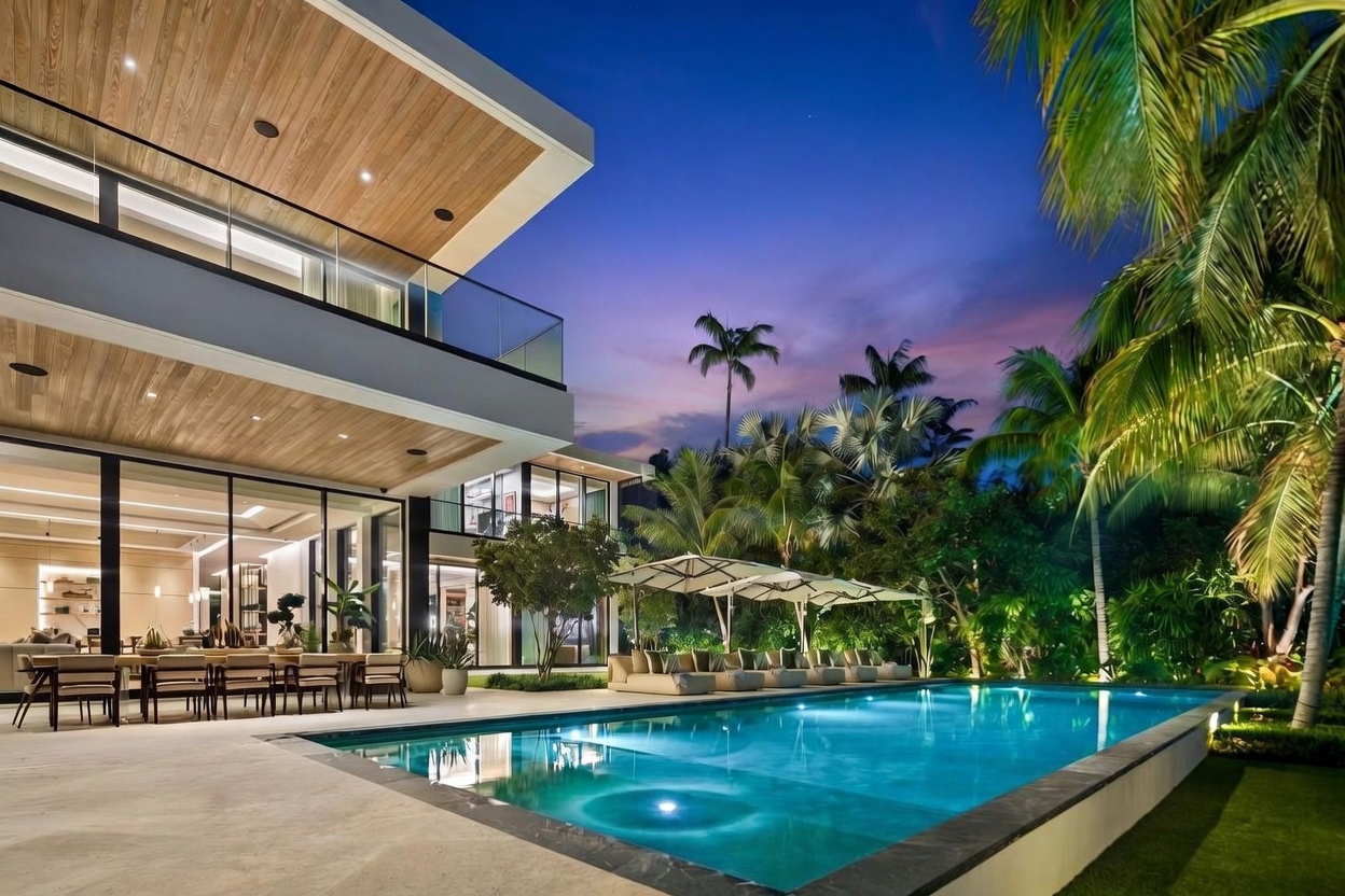 This stunning modern residence features a seamless indoor-outdoor transition with expansive glass walls opening onto a spacious patio and a large, illuminated swimming pool. The exterior showcases clean architectural lines, light wood-paneled ceilings, and a lush, tropical landscape that creates a serene evening atmosphere. The perspective is captured from the poolside, highlighting the luxury outdoor dining area and the sophisticated lighting design of the home.