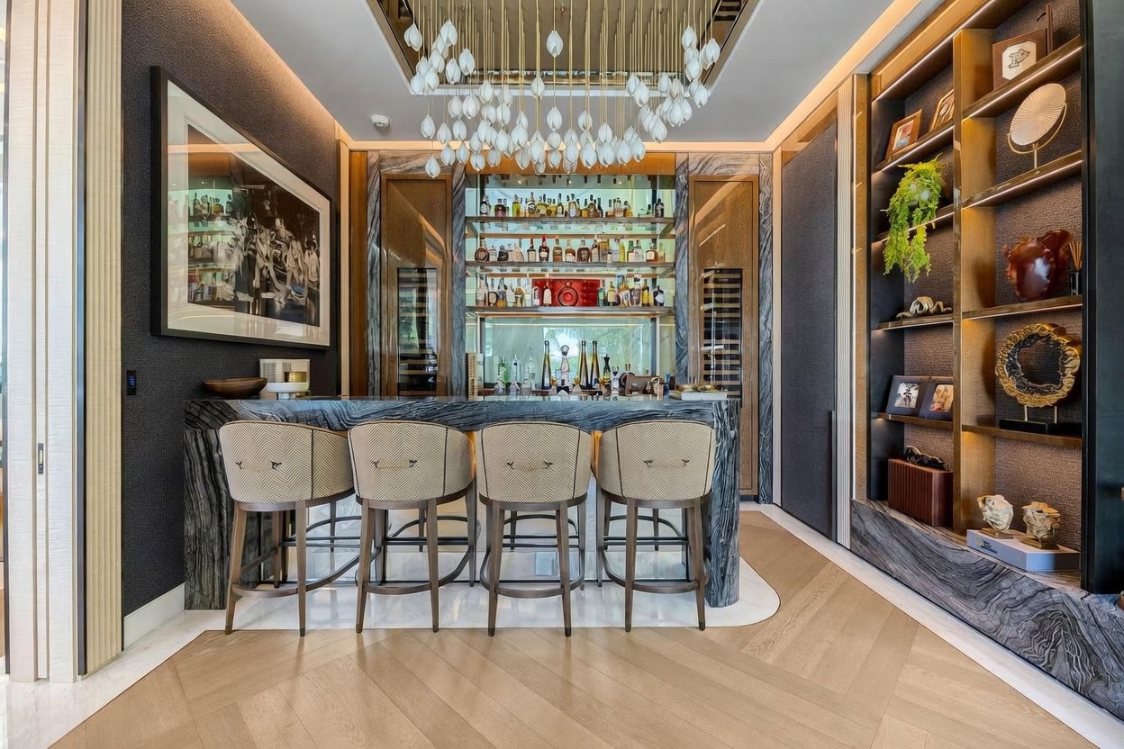 This sophisticated home bar area features a stone-topped counter with four upholstered bar stools, set against a backdrop of dark, textured walls and a mirrored back bar with illuminated shelving. A striking, multi-pendant chandelier hangs from the ceiling, while an adjacent built-in shelving unit displays decorative items and greenery. The space is finished with light-toned wood flooring laid in a herringbone pattern, creating a luxurious and inviting atmosphere.
