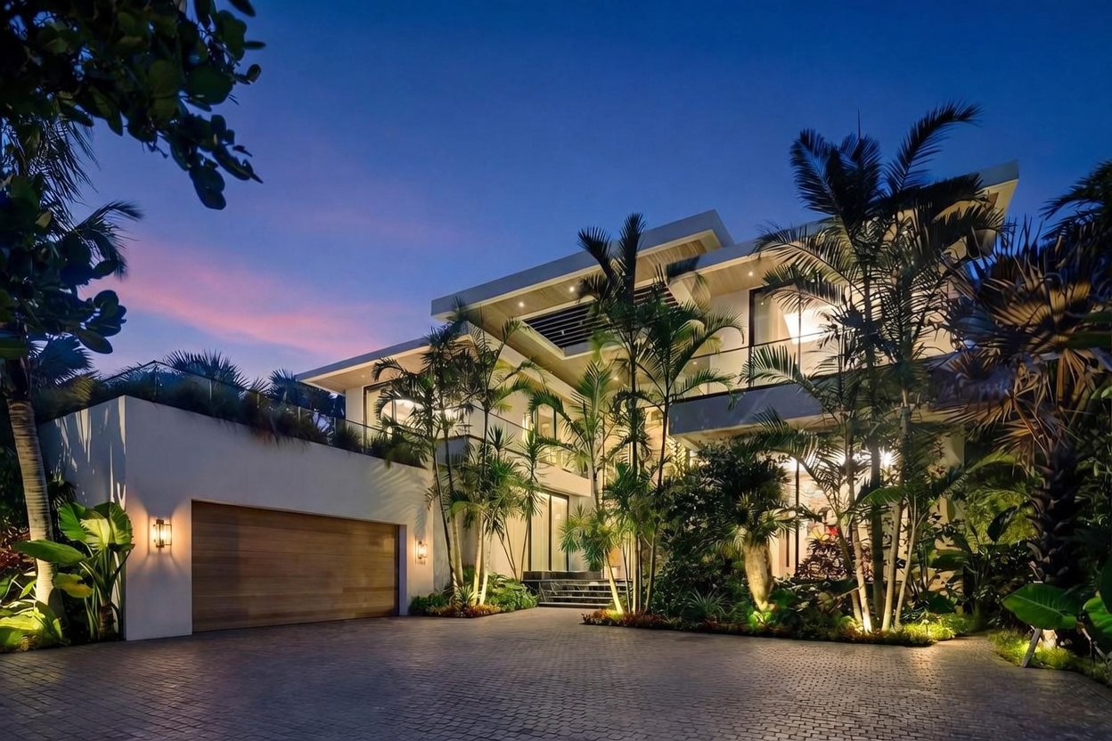 This striking modern residence is captured at dusk, showcasing a multi-level architectural design with clean lines and warm accent lighting. The front facade features a prominent wooden garage door, lush tropical landscaping, and a paved driveway leading to a grand entrance. The cinematic perspective highlights the contrast between the deep blue twilight sky and the inviting glow emanating from the home's interior.