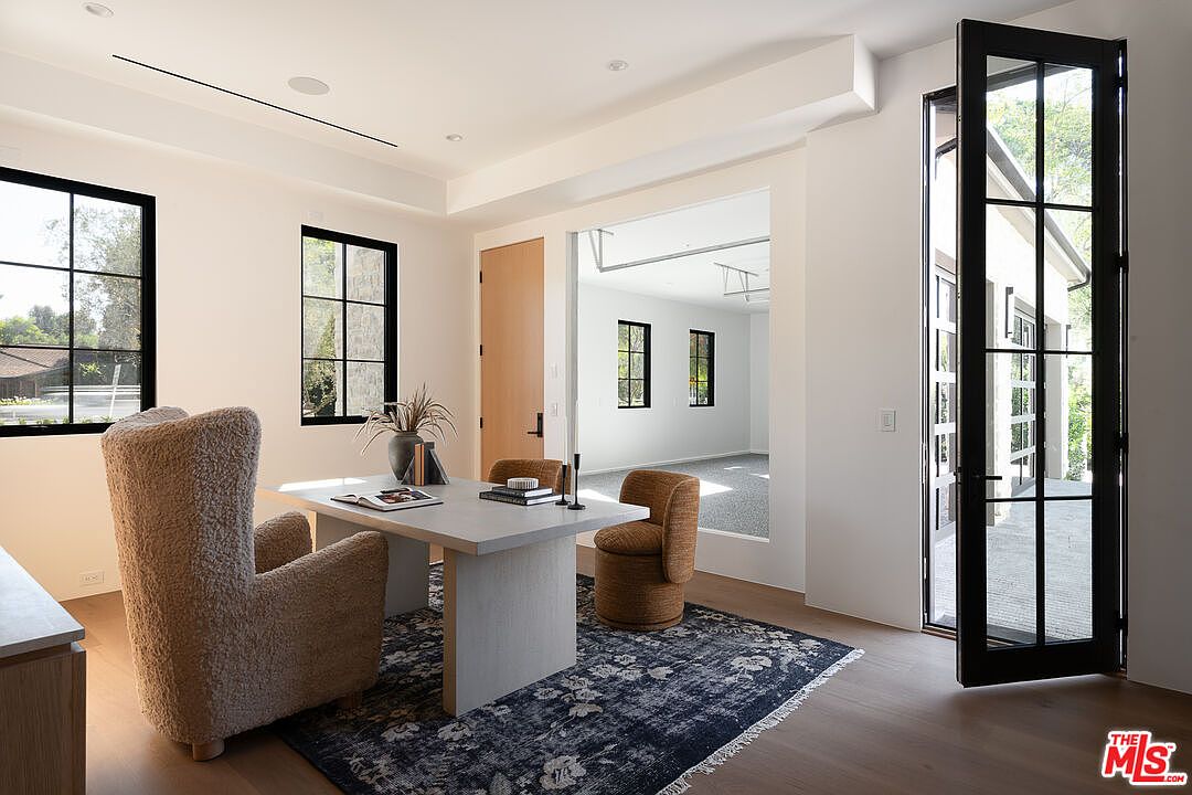 This is an interior shot of a modern office space featuring a large desk with a vase and books, surrounded by stylish chairs on a patterned rug. Natural light streams in through black-framed windows, and an open doorway leads to another room, creating a sense of spaciousness. The overall aesthetic is clean, bright, and conducive to productivity.