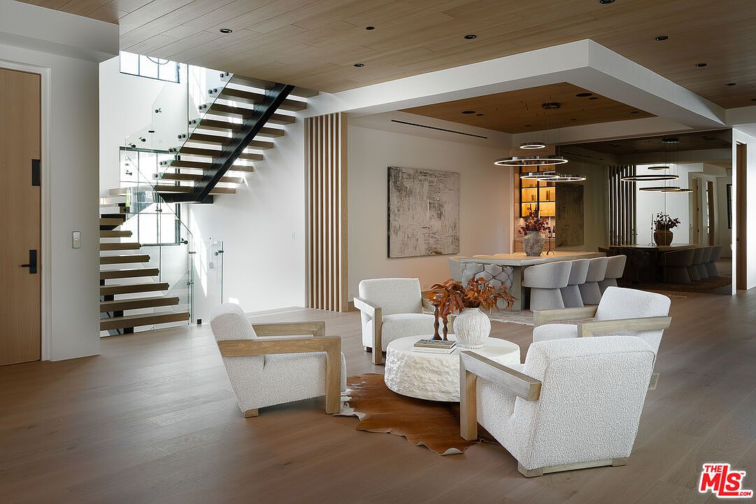 This interior shot showcases a modern living room with an open floor plan, featuring a floating staircase with glass railings and wooden steps. The living area includes four armchairs arranged around a round coffee table on a hide rug, with a view into the dining area with modern lighting fixtures. The room is characterized by its clean lines, neutral color palette, and natural wood accents, creating a sophisticated and inviting atmosphere.
