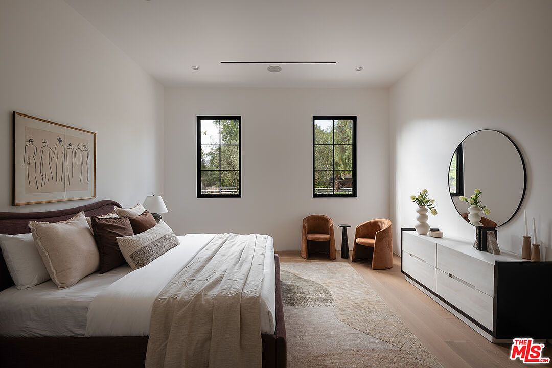 This is a bright and airy primary bedroom featuring a large bed with multiple pillows and a neutral-toned comforter. Two windows provide natural light, and a dresser with a round mirror sits against the far wall. The room is decorated in a minimalist style with a focus on natural materials and textures, creating a serene and inviting atmosphere.