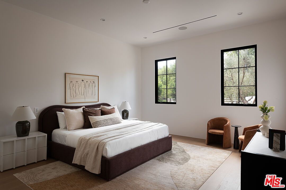 This is a well-lit primary bedroom featuring a large bed with a dark brown frame and a light-colored comforter. The room has two windows with black frames, allowing natural light to fill the space. The walls are painted in a neutral tone, and the room is decorated with artwork, lamps, and two brown chairs, creating a serene and inviting atmosphere.