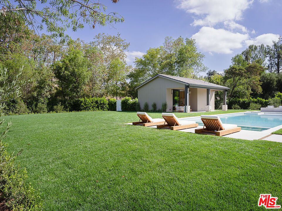This exterior shot showcases a lush green lawn leading to a modern pool house and swimming pool. Three lounge chairs are neatly arranged on a concrete patio beside the pool, inviting relaxation. The scene is framed by mature trees and landscaping, creating a serene and private backyard oasis.