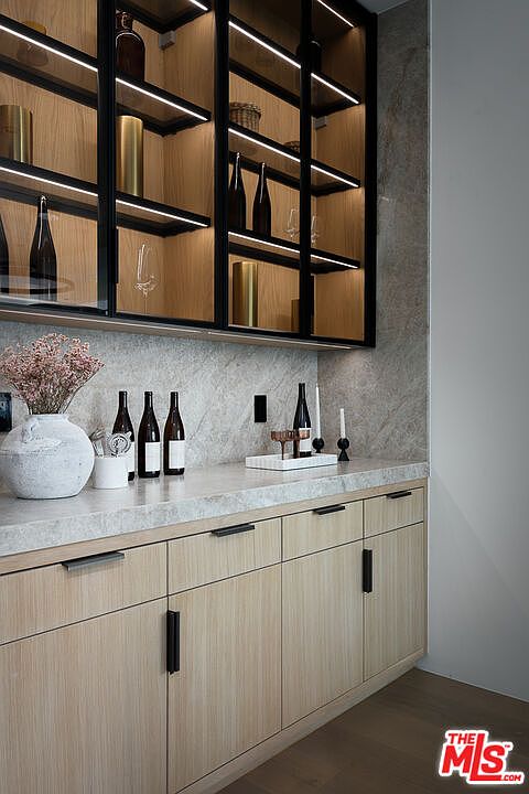 This image showcases a sophisticated wine cellar area, featuring custom light wood cabinetry with sleek black hardware and a marble countertop. The upper cabinets are glass-fronted with integrated lighting, displaying wine bottles and decorative items. The overall design exudes modern elegance and functionality, perfect for wine enthusiasts.