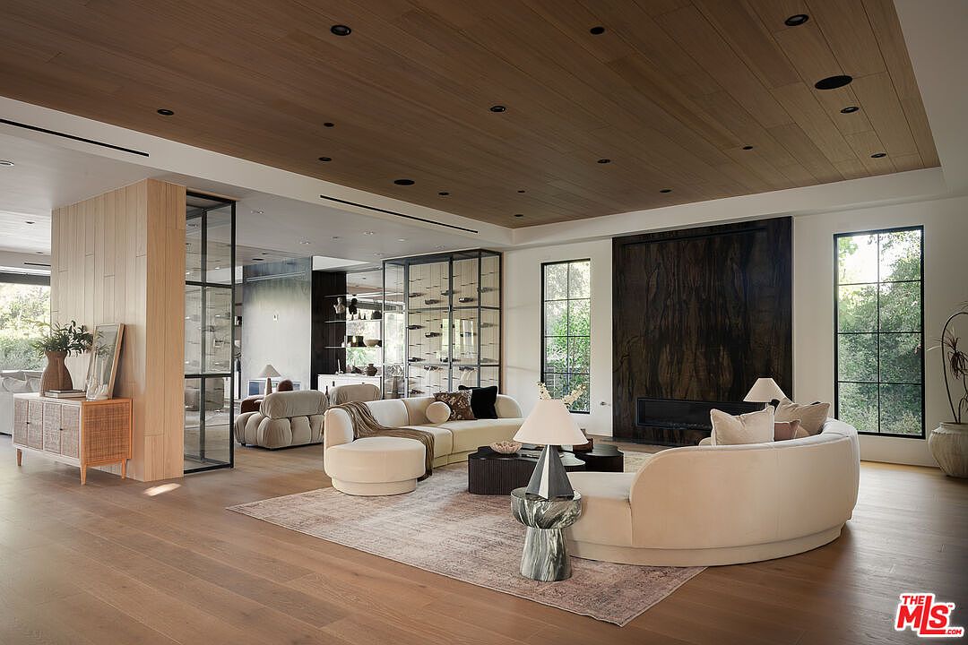 This is a spacious living room featuring a curved sofa, a textured rug, and a wooden ceiling with recessed lighting. Large windows provide ample natural light, and a modern fireplace adds a touch of elegance. The room is decorated in neutral tones, creating a warm and inviting atmosphere.