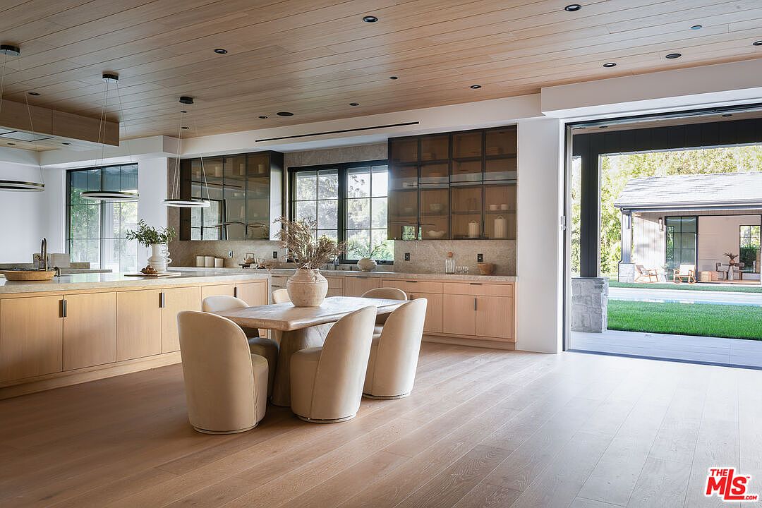 This is a bright and modern kitchen featuring light wood cabinetry, a large island with a stone countertop, and a dining area with four chairs. The kitchen has a seamless indoor-outdoor flow with a large sliding glass door leading to the backyard. The overall impression is one of luxury and sophistication.