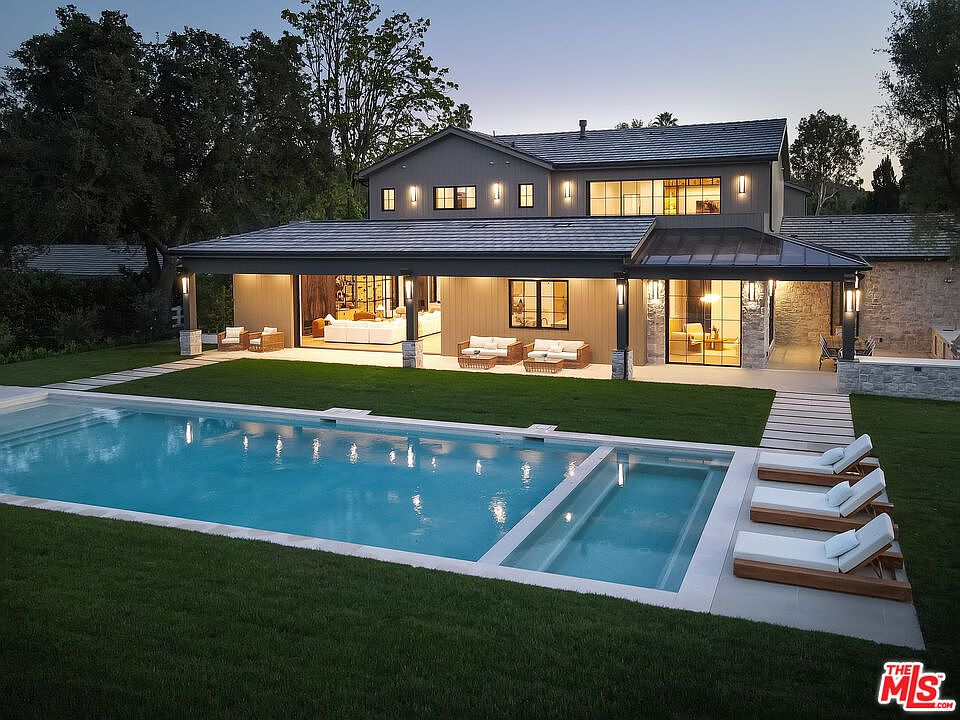 This is a rear exterior view of a luxurious home featuring a rectangular pool with an integrated spa, surrounded by a well-manicured lawn. The house has a covered patio area with outdoor seating, and the architecture is modern with a neutral color palette. The scene evokes a sense of relaxation and upscale living.