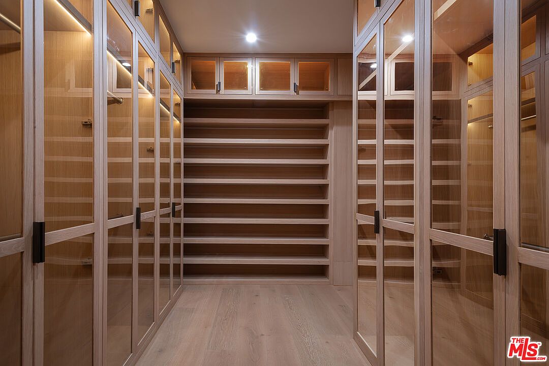 This is a well-organized walk-in closet featuring custom shelving and cabinetry. The closet is illuminated by recessed lighting, and the light wood finish gives it a clean and modern look. Glass doors enclose some of the shelving units, adding a touch of elegance.