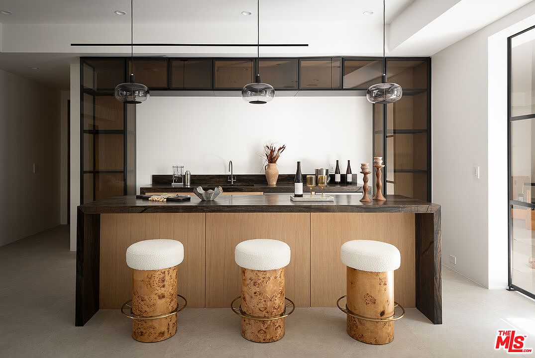 This interior shot showcases a modern kitchen bar area with a dark countertop and light wood cabinetry. Three unique stools with white, fluffy tops and wooden bases with gold accents are positioned in front of the bar. The space is illuminated by pendant lights, and a glass-enclosed shelving unit adds a touch of sophistication.