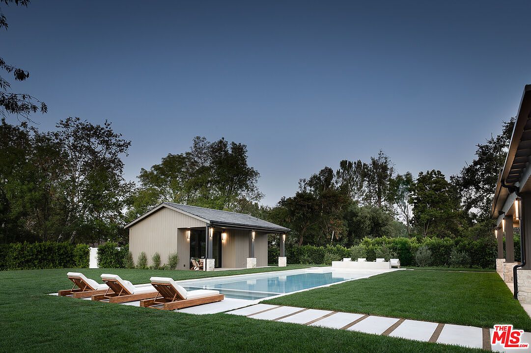 This image showcases a luxurious backyard featuring a rectangular swimming pool with a modern pool house. Three lounge chairs are neatly arranged on the lawn near the pool, inviting relaxation. The well-manicured lawn and stone pathway add to the property's appeal, creating a serene and upscale outdoor living space.