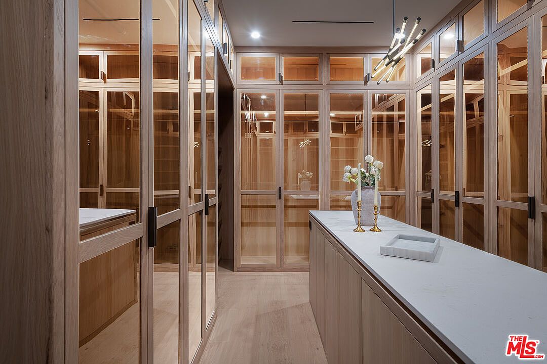 This is an interior shot of a luxurious walk-in closet. The closet features custom wood cabinetry with glass doors, a central island with a white countertop, and modern lighting fixtures. The design is elegant and functional, offering ample storage space and a sophisticated aesthetic.