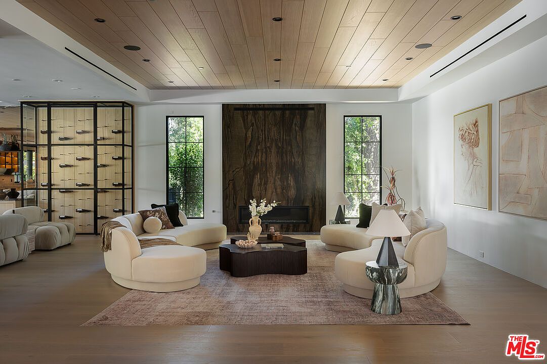 This is a spacious and elegantly designed living room featuring a wooden plank ceiling and hardwood floors. The room is furnished with curved sofas arranged around a unique dark wood coffee table, set upon a patterned area rug. Large windows provide natural light, and artwork adorns the walls, creating a sophisticated and inviting atmosphere.