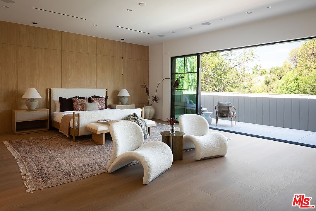 This is a primary bedroom featuring a large bed with a light-colored headboard, flanked by matching nightstands and lamps. The room has a modern aesthetic with wood paneling on the wall behind the bed and large sliding glass doors leading to a balcony. Two white, curved chairs sit in the foreground, adding a touch of contemporary style.