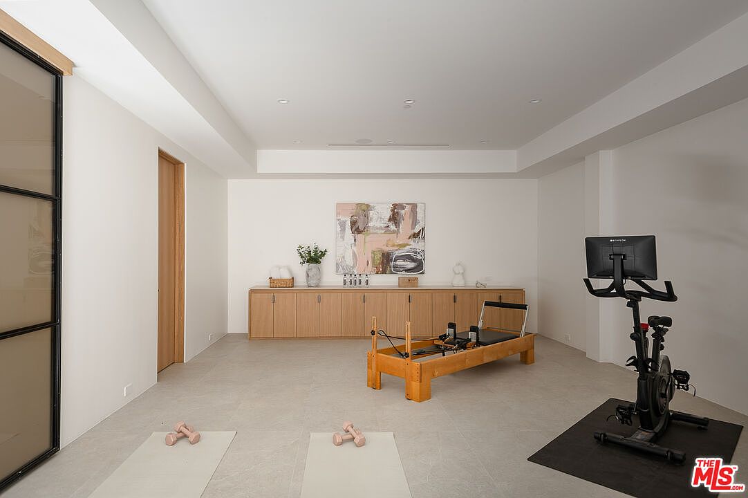 This is a well-lit home gym featuring a Pilates reformer, stationary bike, yoga mats, and dumbbells. A long wooden cabinet lines the back wall, topped with decorative items and a large abstract painting. The room has a clean, minimalist aesthetic with light-colored walls and flooring, creating a spacious and inviting workout environment.