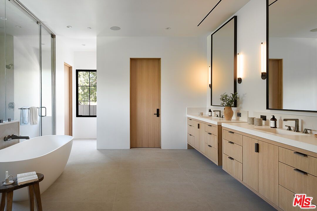 This is a bright and modern primary bathroom featuring a freestanding white bathtub, a double vanity with light wood cabinetry and white countertops, and large mirrors with sconce lighting. The space is clean and minimalist, with neutral-toned tile flooring and a glass-enclosed shower. The perspective is from the doorway, showcasing the entire bathroom layout.