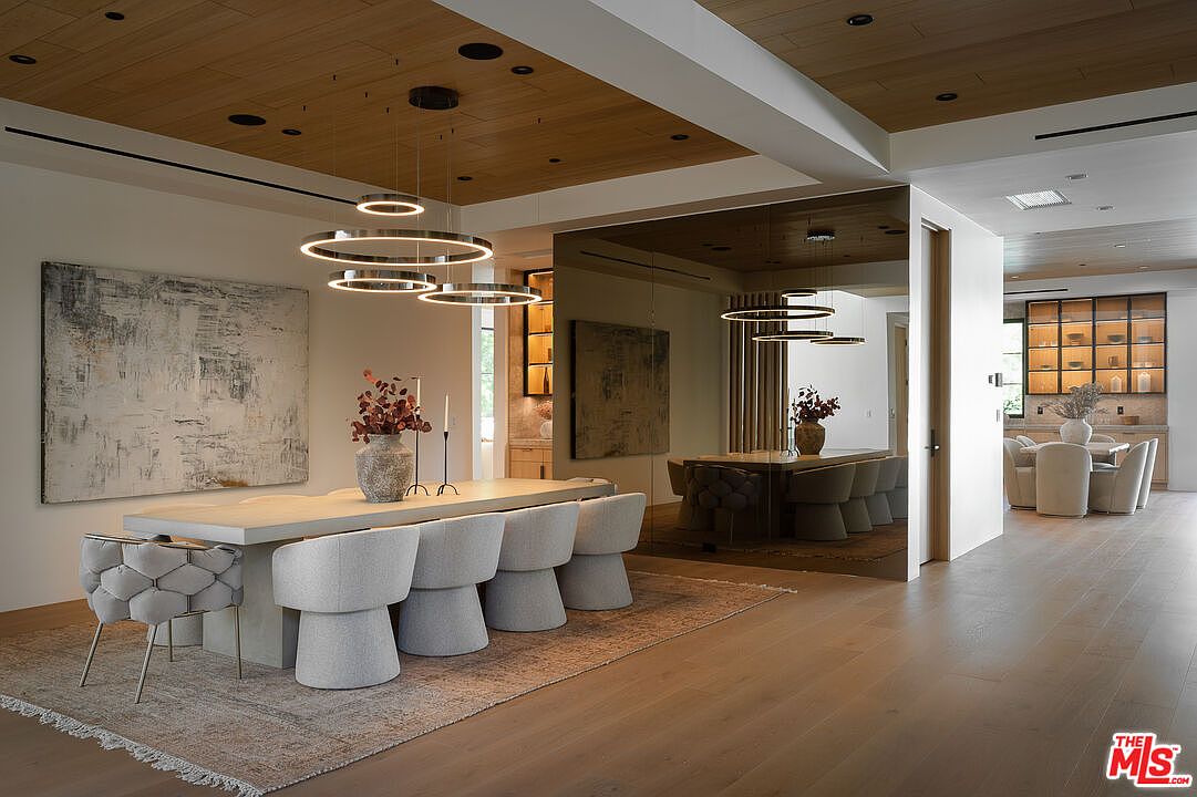 This is an interior shot of a modern dining room featuring a large rectangular table with seating for eight. The room is decorated with a large abstract painting and a unique circular chandelier. The flooring is hardwood, and a large mirror reflects the dining area, creating a sense of spaciousness. The perspective is from the corner of the room, showcasing the table setting and the adjacent room.