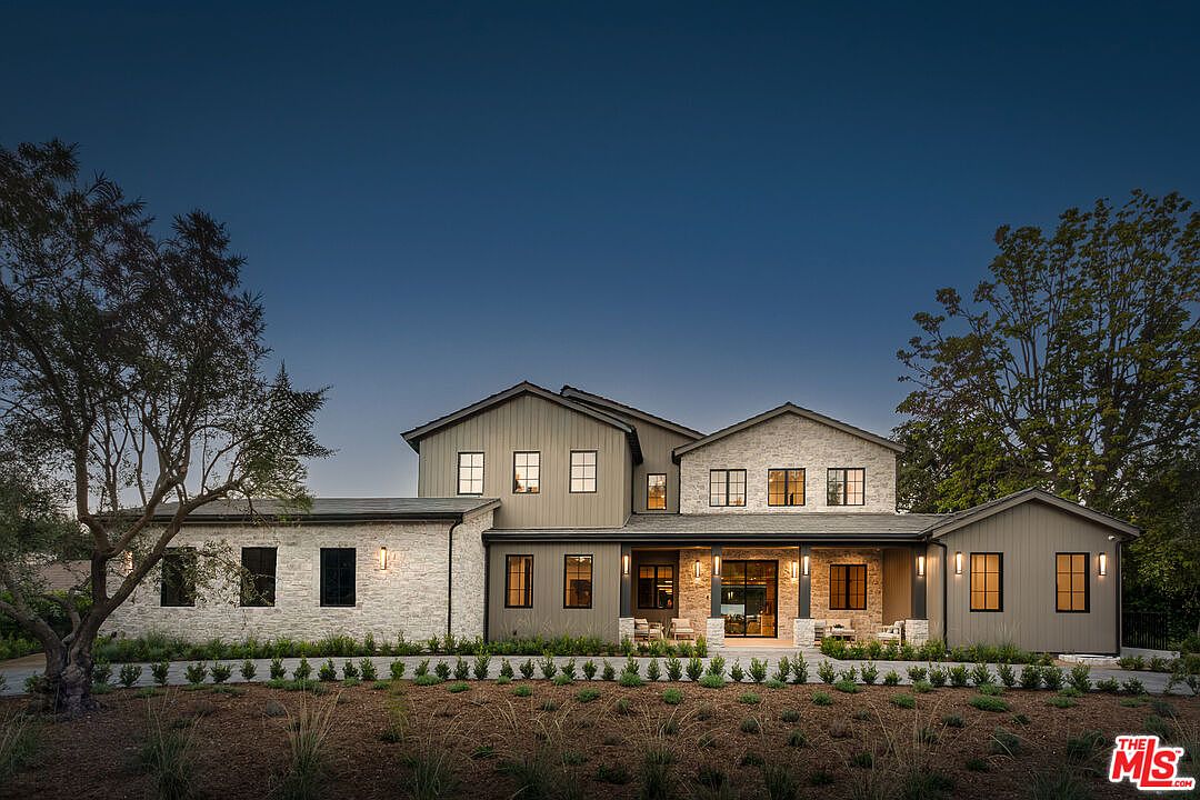 This is a front exterior view of a two-story modern farmhouse-style home at dusk. The house features a combination of stone and siding, with black-framed windows and a covered front porch with stone columns. The landscaping includes manicured shrubs and natural grasses, creating a welcoming and sophisticated curb appeal.