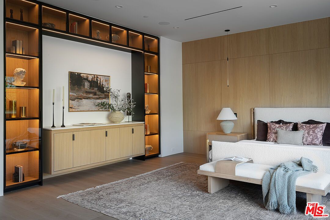 This is a primary bedroom featuring a built-in shelving unit with integrated lighting, a light wood accent wall, and a neutral-toned rug. The bed is adorned with decorative pillows and a throw blanket, and a bench sits at the foot of the bed. The room has a modern and sophisticated aesthetic.