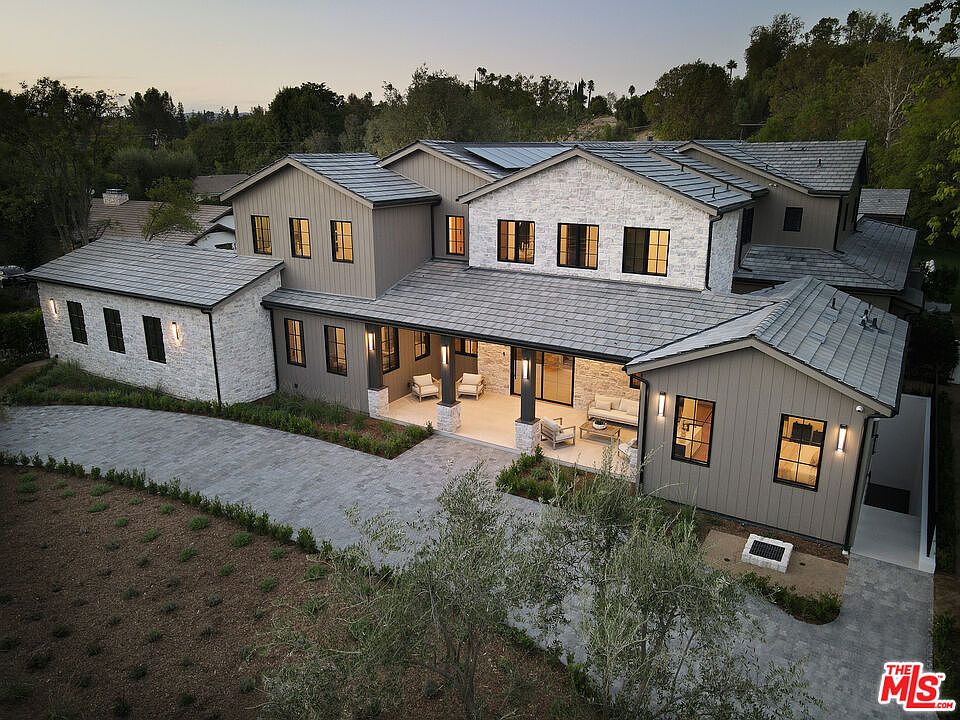This aerial view showcases a sprawling, modern farmhouse-style home with a combination of light stone and gray siding. The roof is a consistent gray tile, and the property features a spacious driveway and landscaping. An inviting covered patio with outdoor seating enhances the home's appeal for outdoor living.