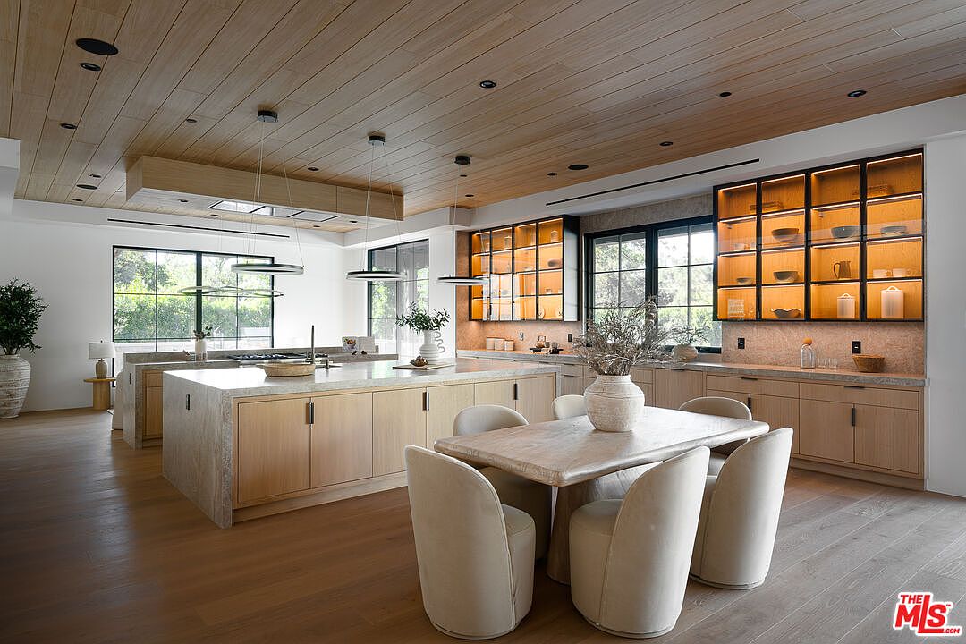 This is a bright and spacious kitchen featuring a large island with a stone countertop and light wood cabinetry. The kitchen also includes a dining area with a stone table and upholstered chairs. The space is illuminated by modern pendant lighting and recessed lighting, with natural light streaming in through large windows, creating a warm and inviting atmosphere.
