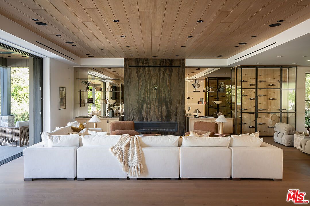 This is a spacious living room featuring a large, modular white sofa as the focal point. The room boasts a modern design with a wooden plank ceiling, a striking stone fireplace, and mirrored built-in shelving units on either side. The hardwood flooring and natural light create a warm and inviting atmosphere, perfect for relaxation and entertaining.