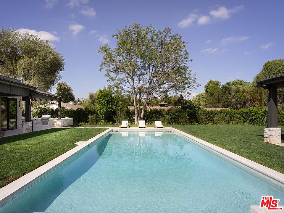 This image showcases a rectangular swimming pool surrounded by a lush green lawn. Three lounge chairs are positioned at the far end of the pool, facing a mature tree that provides shade. The pool's clean lines and the well-maintained landscaping create a serene and luxurious outdoor living space, perfect for relaxation and entertainment.