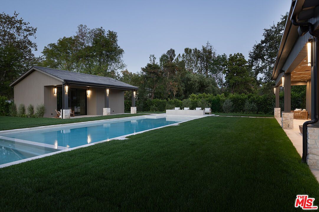 This image showcases a modern backyard with a rectangular pool as the focal point. The pool is surrounded by lush green grass and features a shallow area with lounge chairs. A pool house is visible on one side, and a covered patio area is on the other, creating an inviting outdoor living space.