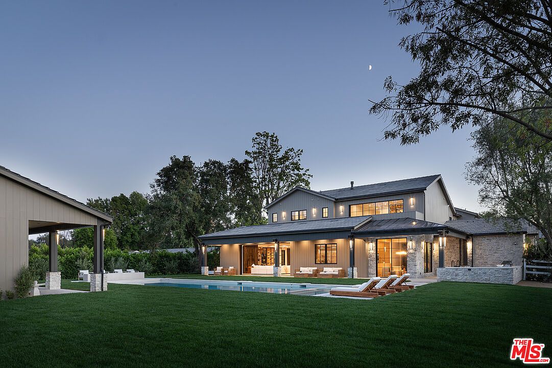 This image showcases the rear exterior of a luxurious home at dusk, featuring a well-manicured lawn, a rectangular swimming pool, and several lounge chairs. The house has a modern farmhouse design with a combination of light-colored siding and stone accents, large windows, and a covered patio area with outdoor seating. The overall impression is one of sophisticated outdoor living and relaxation.