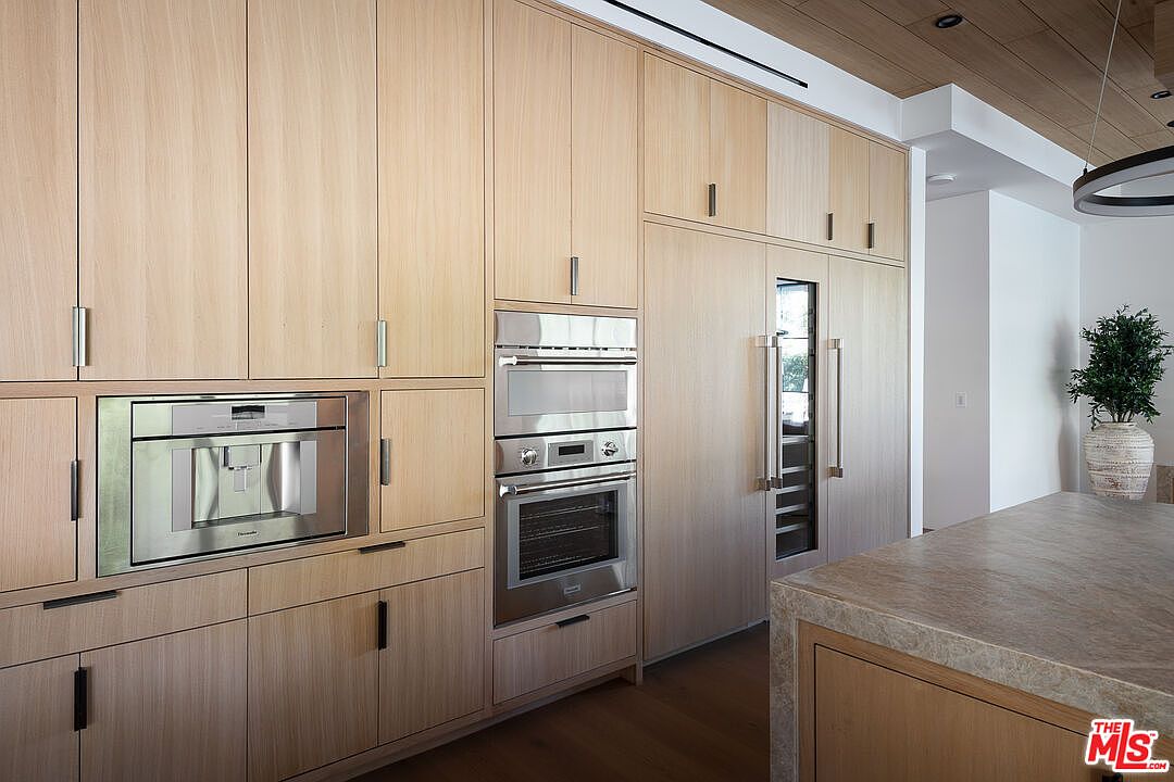 This image showcases a modern kitchen with light wood cabinetry and stainless steel appliances. The built-in appliances include a coffee maker, oven, and refrigerator. The kitchen features clean lines and a minimalist design, creating a sophisticated and functional space.