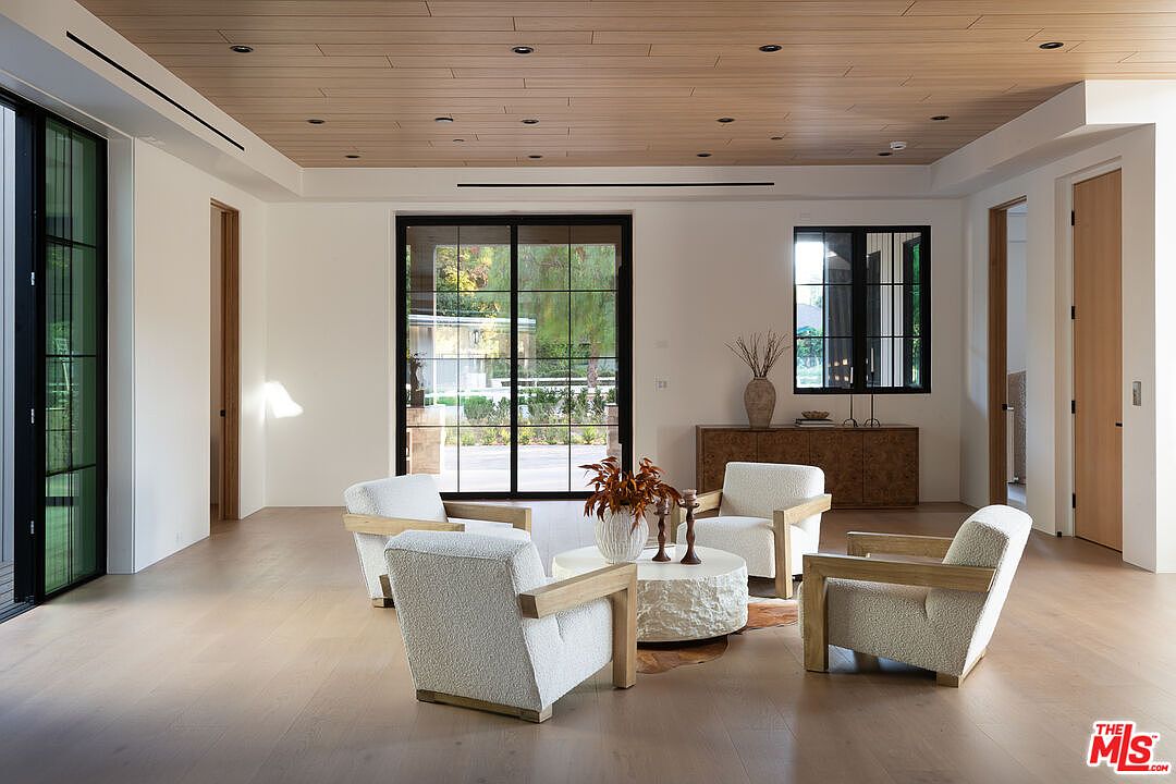 This is an interior shot of a modern living room featuring four armchairs arranged around a round stone coffee table. The room has light wood flooring, a wood plank ceiling, and large windows and doors that provide ample natural light. A wooden cabinet sits against the wall, adding to the room's contemporary and inviting atmosphere.